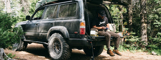 A man on the tailgate of his SUV in the forest.