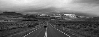 Two guys skateboarding down a lonely highway towards snow capped hills.