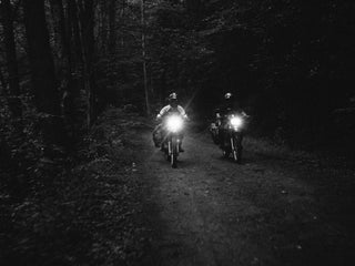 Two motorcyclist riding in the dark down a dirt road in the wilderness.
